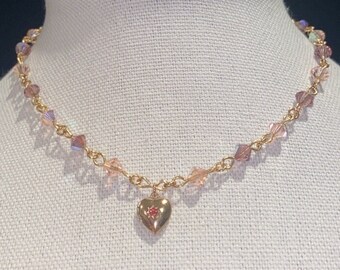 May include: A gold chain necklace with a heart pendant featuring a pink rhinestone. The chain is adorned with small, iridescent pink beads.