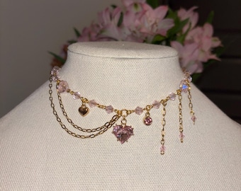 May include: A delicate pink and gold choker necklace. The necklace features a heart-shaped pendant, dangling chains, and pink crystal beads. The necklace is displayed on a mannequin with pink flowers in the background.