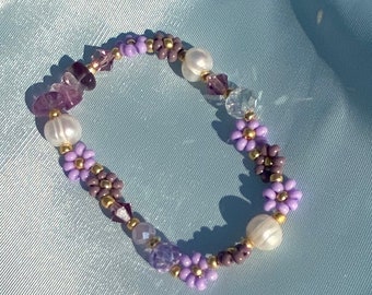 Könnte beinhalten: Ein lila-weißes Armband mit blütenförmigen Perlen und kleinen Kristallperlen.