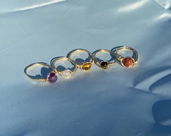 May include: Five gold wire wrapped rings with different colored gemstones. The rings feature amethyst, white, tiger's eye, tiger's eye, and red stones.