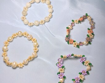 May include: Four beaded bracelets with floral designs. The bracelets are made with white, pink, purple, and green beads. The bracelets are on a light blue background.
