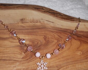 May include: A delicate necklace featuring a gold chain adorned with pink beads and crystals. A snowflake pendant hangs from the center. The necklace is displayed on a wooden surface.