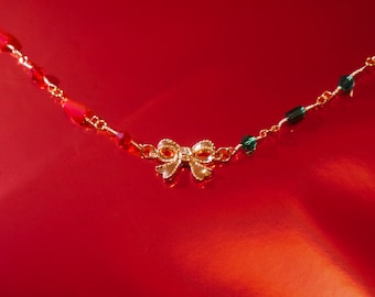 May include: A gold-colored necklace with a bow pendant and red and green beads. The bow is in the center of the necklace, and the beads are strung along the chain. The necklace is on a red background.