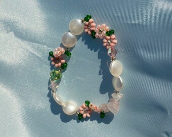 Könnte beinhalten: Ein Perlenarmband mit floralem Design. Das Armband enthält weiße, ovale Perlen, rosa Blumenakzente, grüne Perlen und kleine, klare Kristalle. Das Armband liegt auf einem hellblauen Hintergrund.