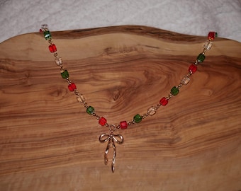 May include: A gold-toned necklace with a bow pendant. The necklace features a chain of red, green, and clear square beads. The bow pendant is a simple, elegant design. The necklace is displayed on a wooden surface.