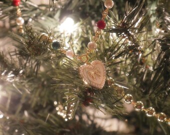 May include: A gold-toned necklace with a heart-shaped pendant featuring a bow design, accented with pearls and red beads, hangs on a Christmas tree. The necklace is set against a backdrop of green pine needles and twinkling lights.