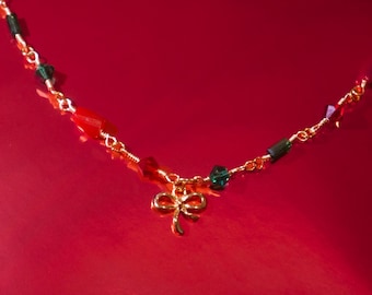 May include: A gold-colored necklace with a bow pendant and a chain adorned with red, green, and clear crystal beads. The necklace is set against a vibrant red background.