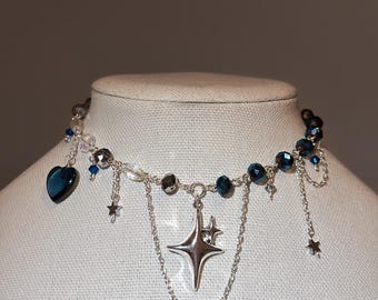 May include: A celestial-themed necklace featuring a silver chain with dangling stars and a four-pointed star pendant. The necklace incorporates blue and clear beads, and a heart-shaped charm. The necklace is displayed on a mannequin.