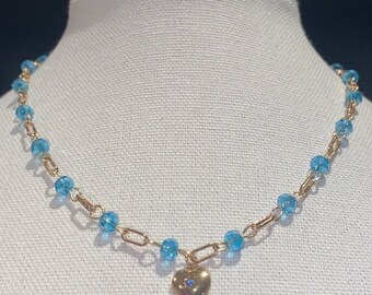 May include: A gold chain necklace with blue glass beads and a small gold heart pendant with a blue stone in the center.
