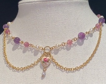 May include: A delicate gold chain necklace with a heart-shaped pendant featuring a floral design. The necklace is layered with a strand of pink and purple beads and white pearls.