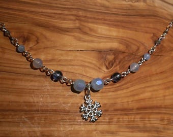 May include: A silver necklace with a snowflake pendant. The necklace features a chain with gray and blue beads. The snowflake pendant is detailed and hangs from the center of the necklace. The background is a wooden surface.