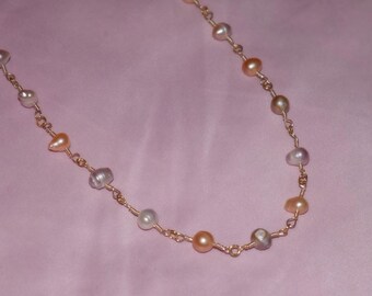 May include: A delicate necklace featuring a gold chain with irregularly shaped, multi-colored pearls. The pearls vary in shades of white, peach, and gray. The necklace is displayed on a pink background, highlighting the jewelry's colors and design.