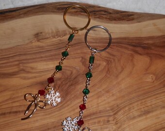 May include: Two Christmas-themed keychains. One is gold-toned, the other silver-toned. Each features a ring, a string of green and red beads, a snowflake charm, and a bow. The keychains are displayed on a wooden surface.