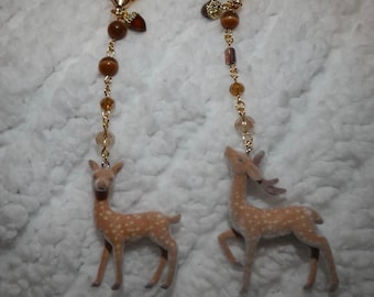 May include: Two keychains with gold-colored heart-shaped clasps and beaded accents. Each keychain features a small, tan-colored deer figurine with white spots. The deer are standing on a white, textured surface.