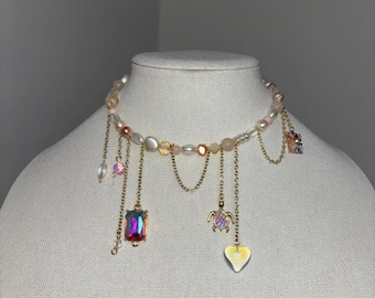 May include: A delicate necklace featuring a strand of pearls and beads in shades of pink and white. Gold chains dangle with iridescent charms, including a rectangular pendant, a turtle, and a heart-shaped charm. The necklace is displayed on a mannequin.