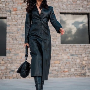 May include: A long black leather coat with a button-up front and a fitted silhouette. The coat is being worn by a person with long dark hair and is standing in front of a stone wall.