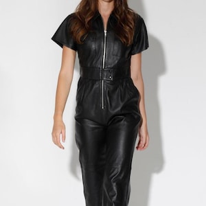 May include: Black leather jumpsuit with short sleeves, a zip-up front, and a belted waist. The jumpsuit features cropped pants and is paired with white sneakers. A modern, stylish outfit.