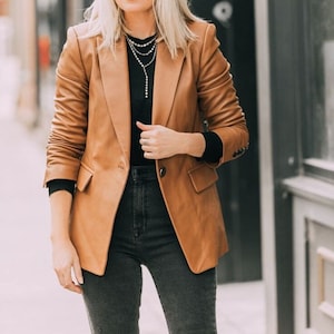 May include: A brown leather blazer with a black mock neck shirt and dark wash jeans. The person is wearing a layered gold necklace.
