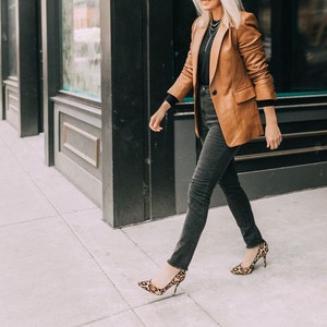 May include: A woman wearing a brown leather blazer, black jeans, and leopard print heels. She is walking towards the camera.