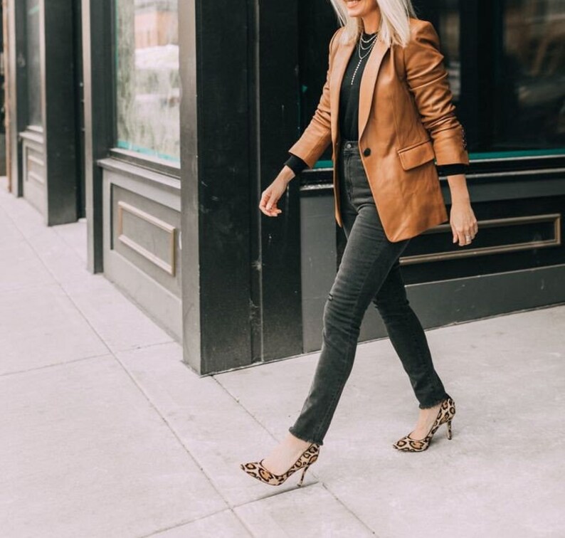 May include: A woman wearing a brown leather blazer, black jeans, and leopard print heels. She is walking towards the camera.