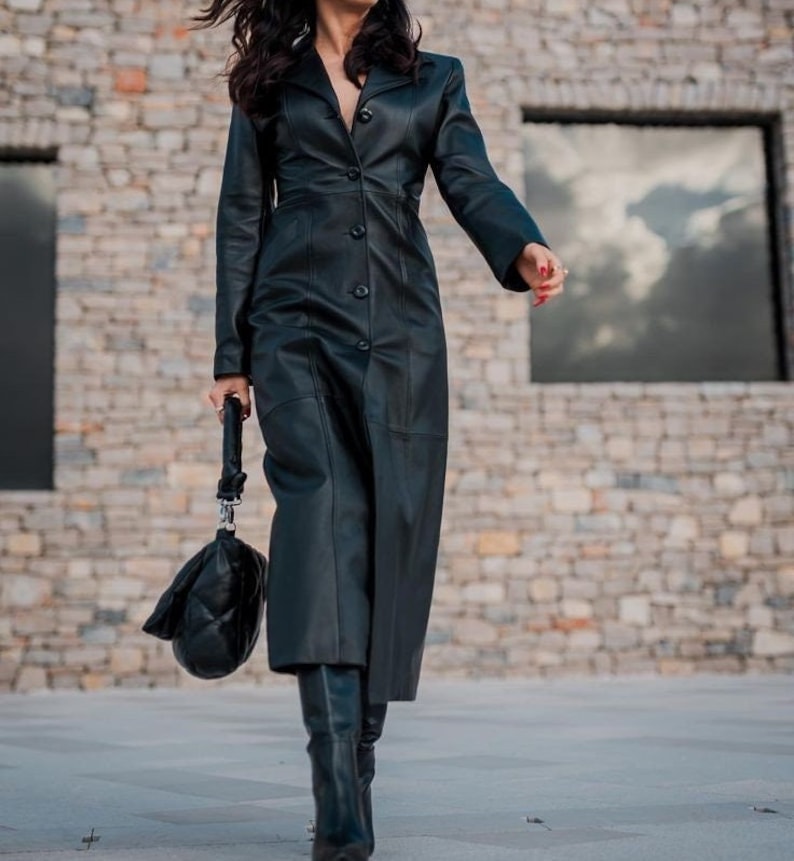May include: A long black leather coat with a button-up front and a fitted silhouette. The coat is being worn by a person with long dark hair and is standing in front of a stone wall.