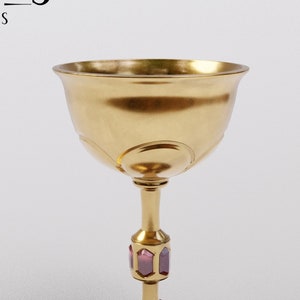 以下が含まれることがあります： 紫色の宝石で飾られた、幅広のボウルと細い茎を持つ金色の聖杯。聖杯の底には、小さく丸い紫色の宝石が飾られています。画像には「PUZZLES AND PROPS」と「PROPS&BEYOND」の文字が含まれています。