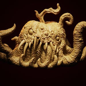 May include: A beige, grotesque sculpture of a creature with multiple eyes, sharp teeth, and tentacles. The sculpture has a textured surface and is set against a dark background. The letters "NS" are in the upper right corner.