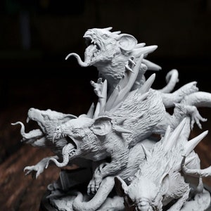 May include: A detailed, grey sculpture of a multi-headed beast with sharp teeth and long tongues, mounted on a black base. A yellow, twenty-sided die is next to the sculpture, suggesting a tabletop game theme.