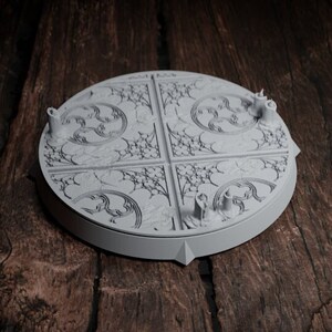 May include: A white, circular, decorative base with a detailed, intricate design. The base has a raised edge and a smooth surface.