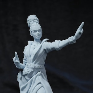 Puede incluir: Una figurita miniatura pintada de blanco de una mujer con un abrigo largo y botas. Está de pie sobre una base redonda con una superficie texturizada. La figurita está en pose con un brazo extendido y la otra mano levantada.