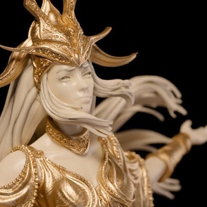 Puede incluir: Una figurita dorada de una mujer con el pelo largo y blanco que lleva una corona y un vestido fluido. La figurita está en pose con los brazos extendidos.