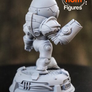 May include: A gray, detailed, 3D printed figurine of a robot with a large head and a single arm extended. The robot is standing on a circular base with a textured surface. The figurine is labeled "Nom Nom Figures."
