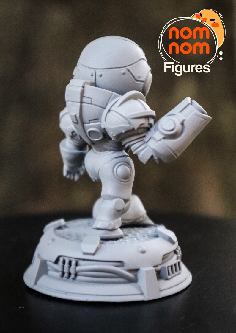 May include: A gray, detailed, 3D printed figurine of a robot with a large head and a single arm extended. The robot is standing on a circular base with a textured surface. The figurine is labeled "Nom Nom Figures."