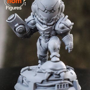 May include: A gray, 3D printed figurine of a video game character, Samus Aran from Metroid, in a chibi style. The figurine is holding a weapon and standing on a base with a detailed design.