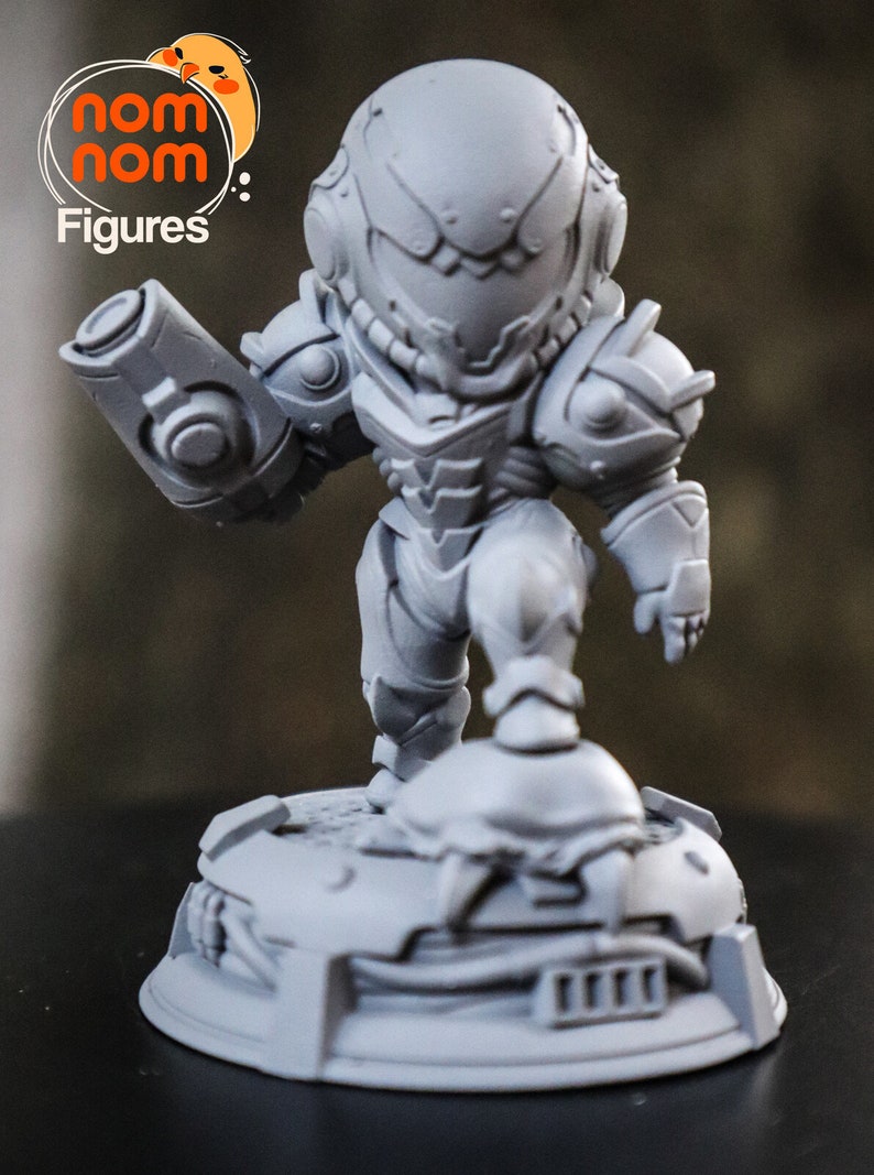 May include: A gray, 3D printed figurine of a video game character, Samus Aran from Metroid, in a chibi style. The figurine is holding a weapon and standing on a base with a detailed design.