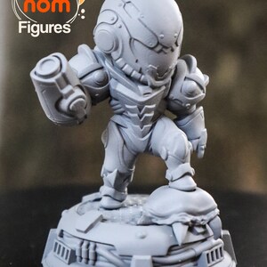 May include: A gray, 3D printed figurine of a video game character, Samus Aran from Metroid, in a standing pose holding a weapon. The figurine is on a base with a textured surface.