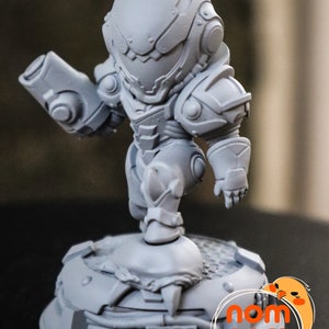 May include: A gray, detailed, 3D printed figurine of a character in a futuristic suit holding a weapon. The figurine is standing on a circular base with a textured surface. The base has the text "nom nom Figures" printed on it.