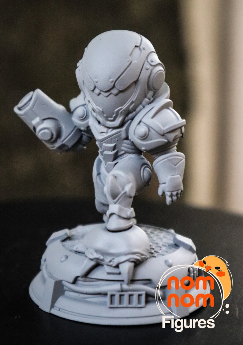 May include: A gray, detailed, 3D printed figurine of a character in a futuristic suit holding a weapon. The figurine is standing on a circular base with a textured surface. The base has the text "nom nom Figures" printed on it.