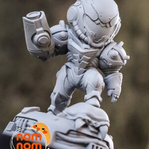 May include: A gray, 3D printed figurine of a space-suited character, standing on a base with the words "nom nom figures" printed on it.