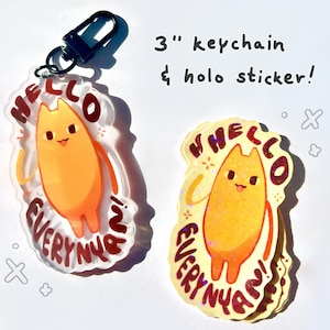 以下が含まれることがあります： 「Hello Everyone!」という赤と白の文字が書かれた、漫画の猫が描かれた7.6cmのキーホルダーとホログラフィックステッカー。猫はオレンジ色で、ピンクの舌を出しています。