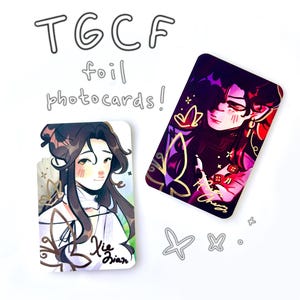 Puede incluir: Dos tarjetas fotográficas rectangulares con acabado metalizado e ilustraciones estilo anime. Una tarjeta muestra un personaje de pelo largo oscuro etiquetado como Xie Lian, la otra un personaje en tonos oscuros. Texto: TGCF foil photocards! y mariposas.