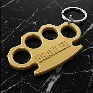 May include: Gold-colored keychain featuring a brass knuckle design with three finger holes. The word "BRAWLER" is embossed on the front. The keychain has a silver key ring attached.