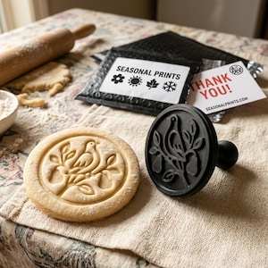 May include: A black circular cookie stamp featuring a bird perched on a branch with leaves and pears, alongside a stamped cookie showing the same design. A black bubble mailer labeled SEASONAL PRINTS and a white THANK YOU card are visible.