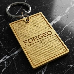 May include: A gold-colored rectangular keychain with a woven texture. The word "FORGED" is engraved across the top. The keychain is attached to a silver metal ring. The background is a black and white marble surface.