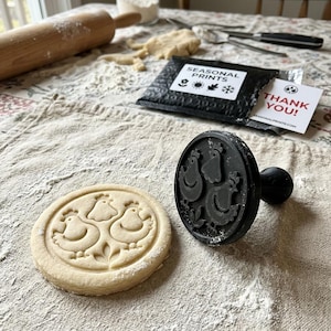 May include: A black circular cookie stamp with a chicken design and a matching round dough piece featuring an embossed chicken pattern. The stamp has a handle, and the dough rests on a floured cloth near a black envelope labeled SEASONAL PRINTS.
