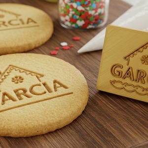 May include: A wooden cookie stamp with a house design and the word "Garcia" engraved on it. The stamp is on a wooden surface next to two cookies with the same design.