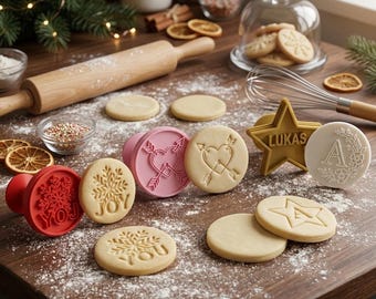 May include: A collection of round and star-shaped cookie stamps and cutters made of plastic. The stamps feature designs including a snowflake with the text JOY, a heart with arrows, the name LUKAS, and the letter A with floral accents.
