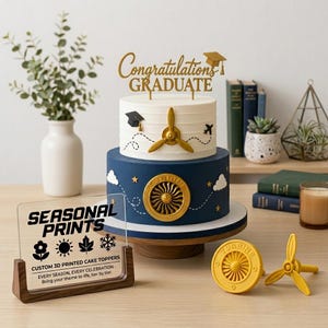 May include: A two-tiered cake featuring a white top layer with a gold airplane propeller and graduation cap, and a navy blue bottom layer with a gold turbine emblem. A gold "Congratulations GRADUATE" topper sits on top. Includes a sign reading "SEASONAL PRINTS".