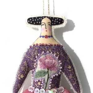 May include: A handmade fabric doll with a floral design. The doll features a purple dress with a pink rose, a pink skirt, and a black and white hat. The doll is adorned with beads and sequins and has a gold string for hanging.