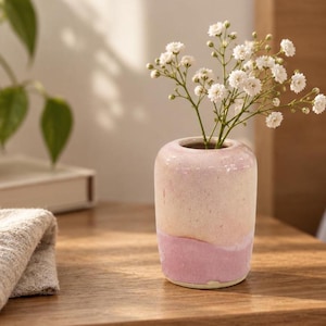May include: A small, cylindrical ceramic vase with a smooth, glossy finish featuring a two-tone colour scheme of pale cream on the upper portion and a soft, muted pink on the lower section, containing a sprig of small white baby's breath flowers.
